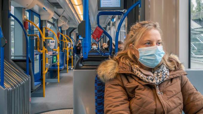 A woman with shoulder-length blonde hair is on a public transit train. She is wearing a brown coat with a fur-trimmed hood, a scarf and a blue surgical mask.