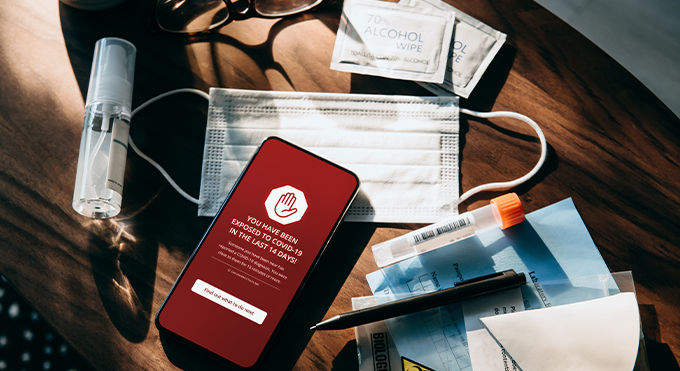 A smartphone is shown with a red screen and a warning that you have been exposed to COVID-19 within the last 14 days. The phone is sitting diagonally tilted about 30 degrees to the right on a surgical mask and surrounded by two packets of alcohol wipes at about 1:00 o'clock, a plastic laboratory specimen collection vial with a bright orange top at about 4:00 o'clock also laying at about an 80 degree angle , a black pen just below sits at angle parallel to the specimen vial on top of a light blue paper that has the word "laboratory" visible, a clear bottle of sanitizer spray lays nearly vertically at 9 o'clock and a pair of tortoiseshell glasses sits folded at about 11 o'clock.