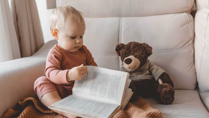 A baby sits on a white couch reading a book next to a teddy bear.
