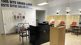 Open room with drug consumption booths, crash carts and text on the back wall that reads "This site saves lives" in English and Spanish.