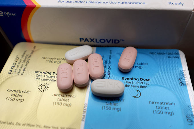 Six oval-shaped pills rest on yellow and blue packaging. A white box that says "Paxlovid" is in the background.
