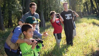 In the foreground, Trish O’Kane points and guides a student holding binoculars amid a forest. More students stand in the background and look into the distance, some through binoculars.