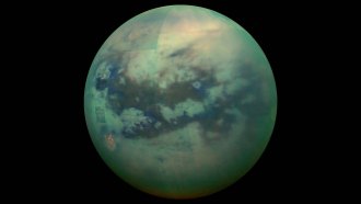 Infrared composite image of Titan, Saturn's largest moon. The moon appears blue-green in the image with dark splotches across its surface.